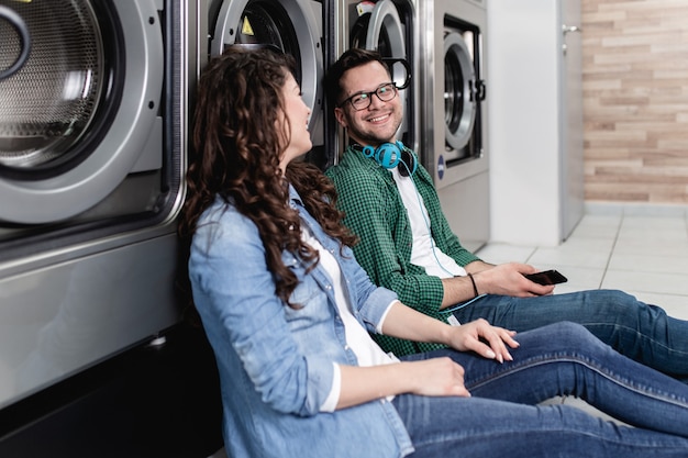 young cheerful couple doing laundry together at laundromat shop.