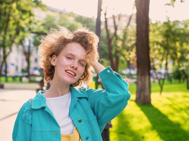 Photo young cheerful curly redhead woman in blue jacket holding straw hat in her hands and happy