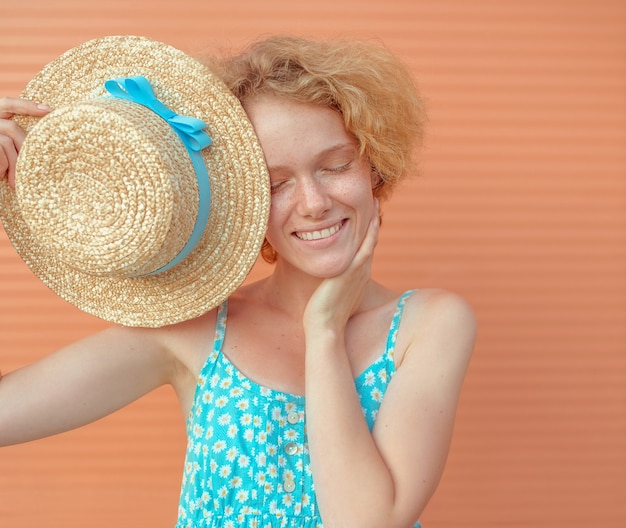 young cheerful curly redhead woman in blue sundress holding straw hat in her hand on beige backgroun