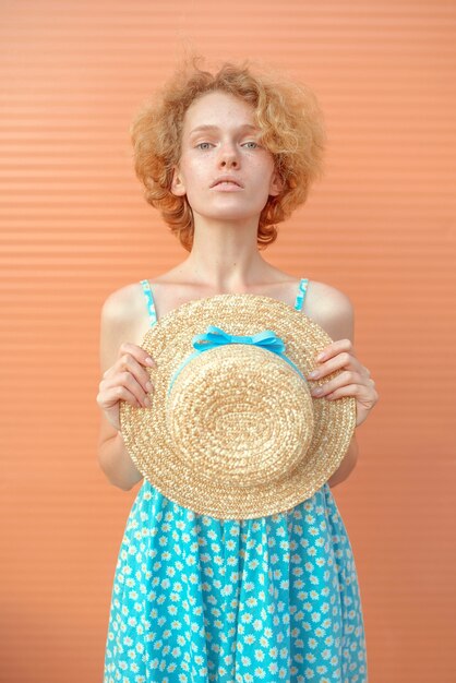 young cheerful curly redhead woman in blue sundress holding straw hat in her hand on beige