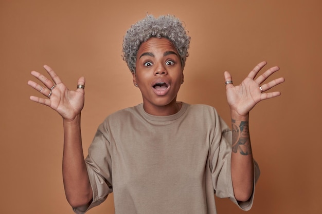 Young cheerful ethnic African American woman with big eyes raises hands up overjoyed rejoicing in own achievements and dreams come true standing in brown studio Achievement success concept