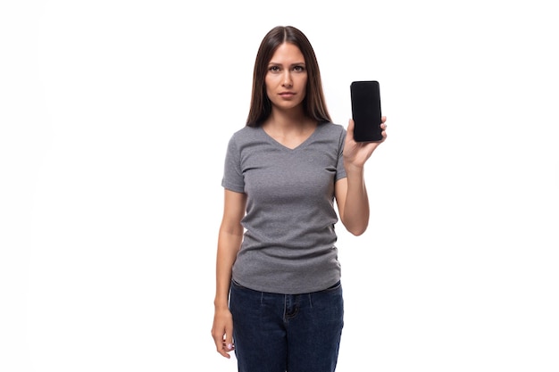 Young cheerful european brunette woman dressed in gray basic tshirt holding smartphone with screen