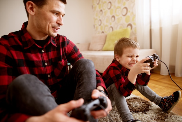 Young cheerful excited father and son in the same red shirt playing console games with gamepads in a bright living room.