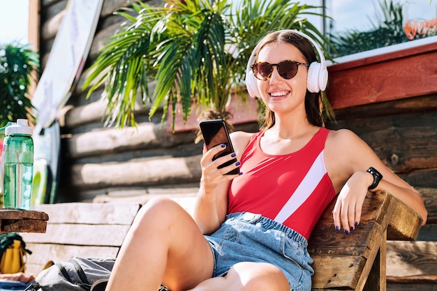 Young cheerful female in headphones, sunglasses, red tanktop and denim shorts sitting on wooden bench and listening to music at resort