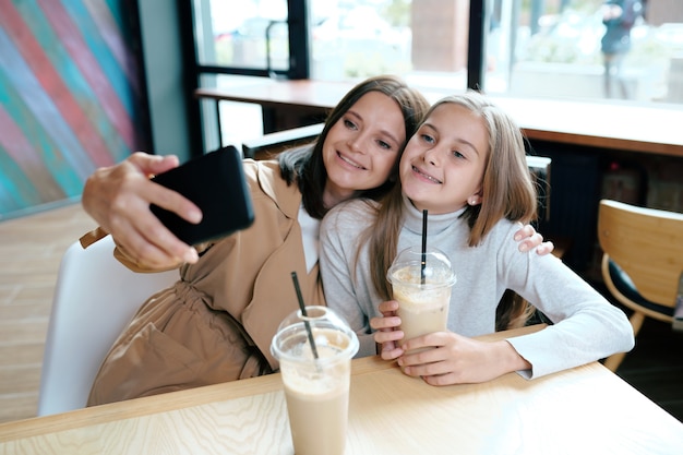 Giovane femmina allegra e sua figlia in abbigliamento casual che fa selfie mentre riposa nella caffetteria e sorseggiando gustose bevande