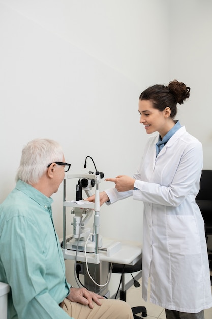 Photo young cheerful female ophthalmologist in whitecoat standing by medical equipment in clinics while going to test eyesight of senior male patient