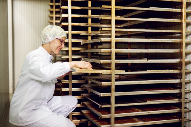 Young cheerful female worker in a sterile cloth examining freshly baked cookies on a cookie shelf in bakery storage room.