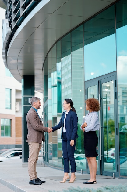 Young cheerful and friendly businesswoman welcoming new colleague by handshake while meeting him by entrance of office building