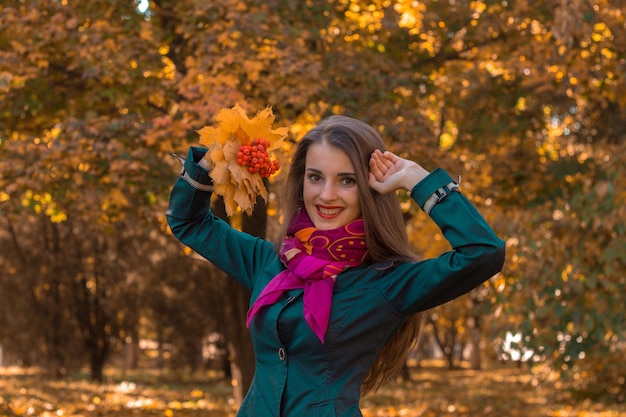 Молодая жизнерадостная девушка в розовом платке подняла руки к голове, держит листья и улыбается