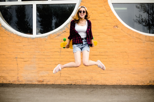 Photo young cheerful girl posing with yellow skateboard against orange wall.