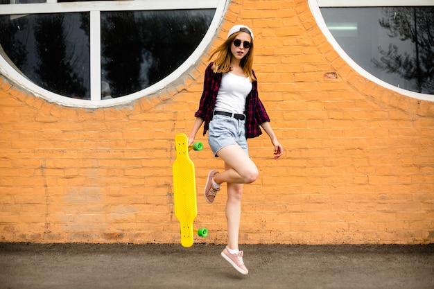 Young cheerful girl posing with yellow skateboard against orange wall.