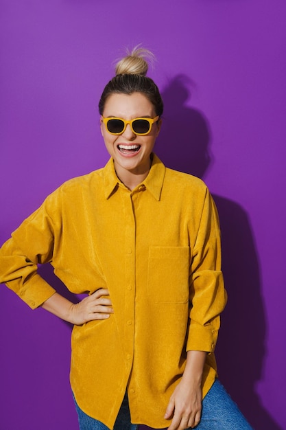 Young cheerful girl wearing yellow shirt and sunglasses against purple background