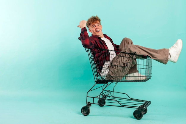 Photo young cheerful guy sits in shopping cart and points back on blue background