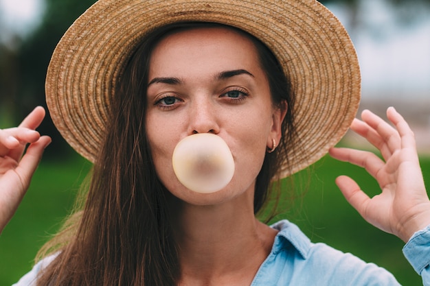 Young cheerful happy hipster woman in hat blowing bubble of chewing gum outdoor