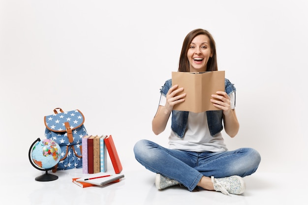 Giovane studentessa felice allegra in vestiti di denim che tiene la lettura del libro seduta vicino al globo, zaino, libri di scuola isolati