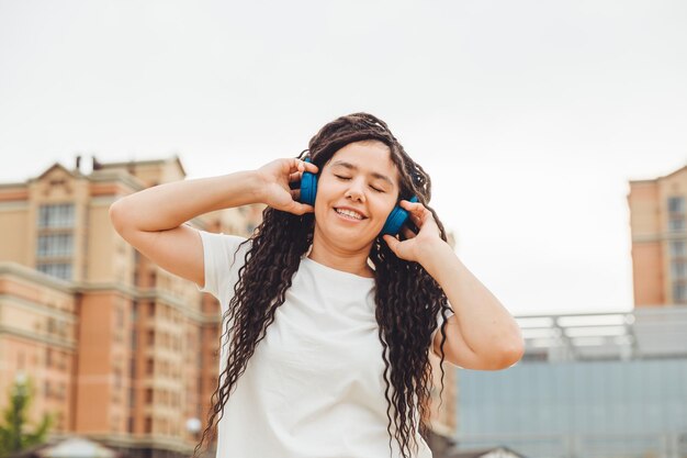 Foto una giovane donna allegra e felice con i dreadlocks vestita con una maglietta bianca che balla ascoltando musica con le cuffie che riposano rilassandosi in un parco cittadino camminando lungo un vicolo concetto di stile di vita urbano