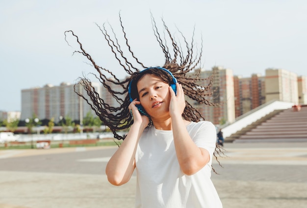 白い t シャツに身を包んだドレッドヘアを持つ若い陽気な幸せな女性は、路地に沿って歩く都市公園でリラックスして休んでヘッドフォンで音楽を聴いて踊る白い t シャツ アーバン ライフ スタイル コンセプト