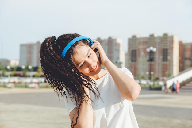 白い t シャツに身を包んだドレッドヘアを持つ若い陽気な幸せな女性は、路地に沿って歩く都市公園でリラックスして休んでヘッドフォンで音楽を聴いて踊る白い t シャツ アーバン ライフ スタイル コンセプト