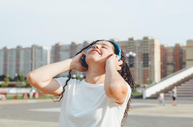 A young cheerful happy woman with dreadlocks dressed in a white Tshirt dancing listening to music with headphones resting relaxing in a city park walking along an alley Urban lifestyle concept