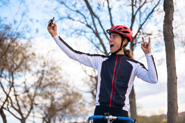 Молодой жизнерадостный мужской велосипедист в спортивной одежде и шлеме показывая знак руки и принимая selfie на smartphone в парке