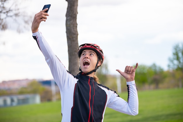 Молодой жизнерадостный мужской велосипедист в спортивной одежде и шлеме показывая знак руки shaka и принимая selfie на smartphone в парке