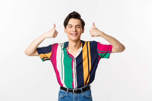 Young cheerful man smiling, showing thumbs up in approval, praise something good, standing over white.