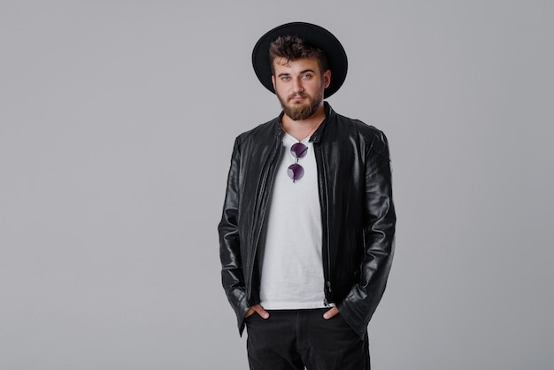 A young cheerful man with a beard in a stylish black leather jacket and hat on a gray wall	