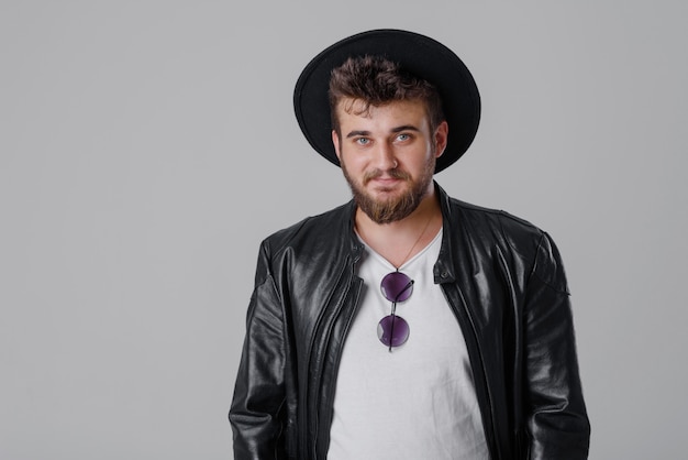 A young cheerful man with a beard in a stylish black leather jacket and hat on a gray wall