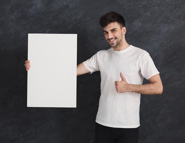 Young cheerful man with blank white banner and recommend. Smiling man holding advertising sheet and showing thumb up gesture, gray studio background, copy space