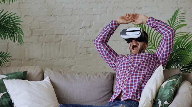 Young cheerful manhaving 360 VR video experience while sitting on couch in living room at home