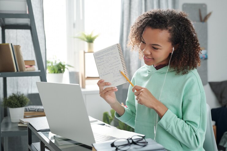 Photo young cheerful mixed-race female student pointing at notes on page of copybook while explaining them to her friend on laptop display