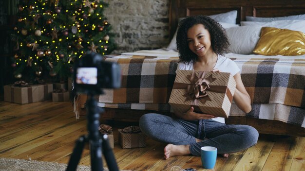 Young cheerful mixed race girl recording video blog about packing christmas gift box at home