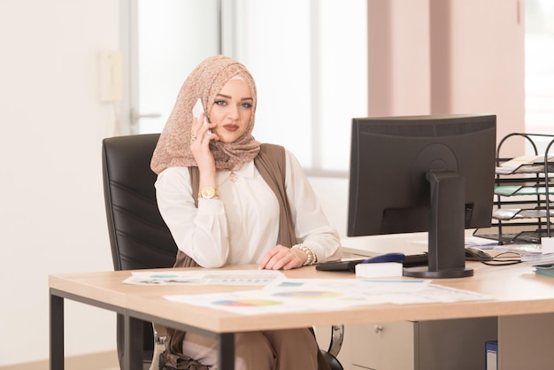Молодая веселая мусульманская деловая женщина, работающая с компьютером за столом в современном офисе и разговаривающая по телефону