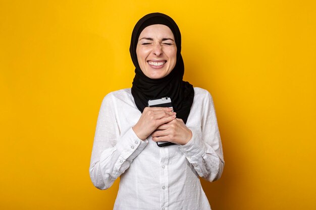 Young cheerful muslim woman in hijab holds a phone in her hands and rejoices or celebrates cheto something on a yellow background banner