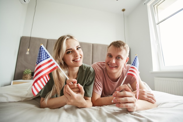 Young cheerful patriots with American flags lying on bed