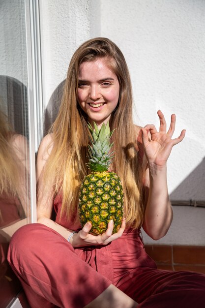 Young cheerful sensual woman in casual summer dress holding tropical pineapple with closed eyes and showing ok gesture