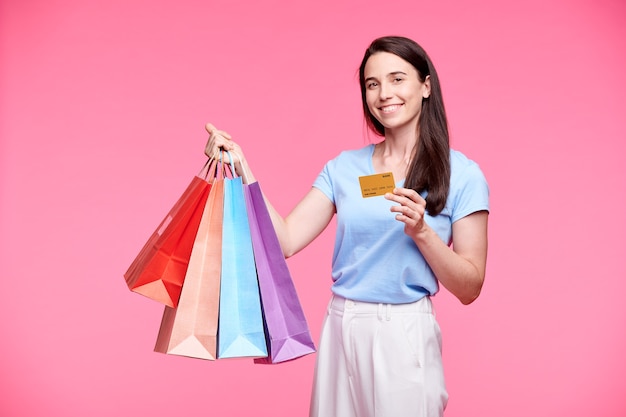 Young cheerful shopper with several paperbags showing plastic card which she used for paying for goods