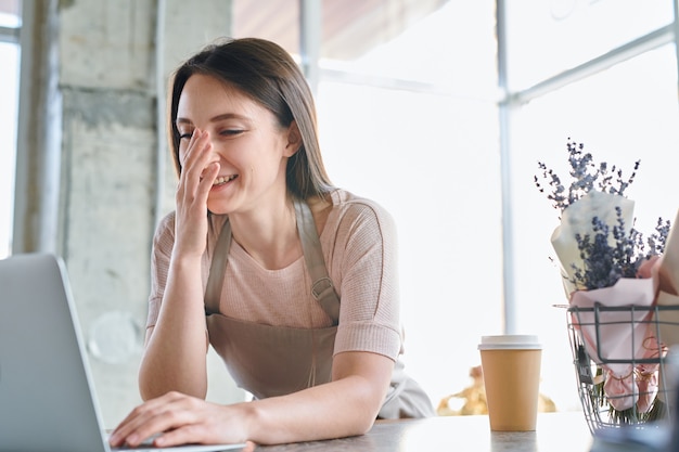 Young cheerful or slightly confused florist looking at laptop display with hand by her face during work