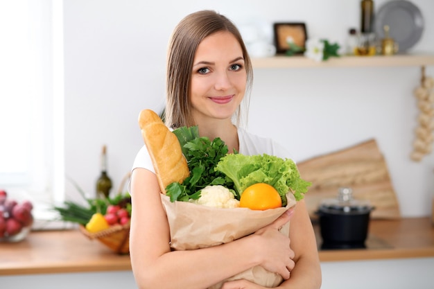 若い陽気な笑顔の女性は、キッチンで料理をする準備ができています主婦は新鮮な野菜や果物でいっぱいの大きな紙袋を持ってカメラを見ています