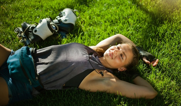 公園の芝生の上に横たわって休んで、ヘッドフォンで音楽を聴く若い陽気なスポーツ女性ローラースケート選手
