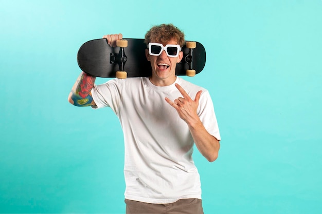 young cheerful tattooed guy in white tshirt holds skateboard and shouts on blue isolated background