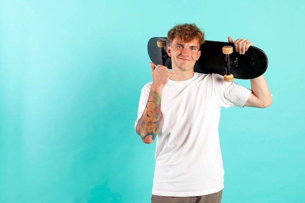 Young cheerful tattooed guy in white tshirt holds skateboard and smiles on blue background