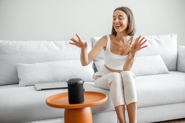 Young and cheerful woman controlling home devices with a smart speaker, sitting on the couch at home. Smart home and managing home by a voice commands concept