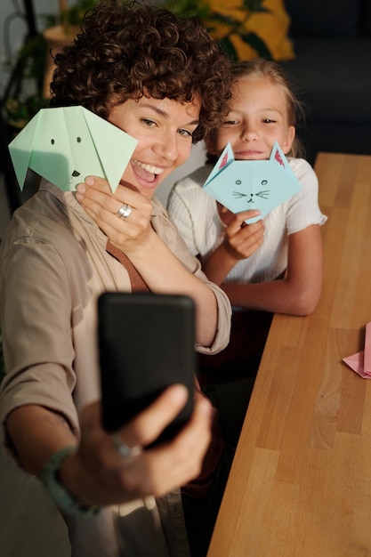 Giovane donna allegra e sua figlia con origami che fanno selfie al tavolo