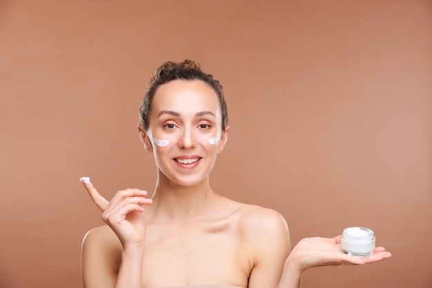 Photo young cheerful woman holding small jar with moisturizing facial cream on palm while applying it after morning or evening hygiene