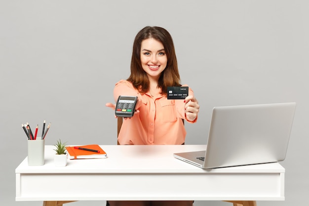 Young cheerful woman holding wireless modern bank payment terminal to process acquire credit card payments work at desk with pc laptop isolated on gray background. Achievement business career concept.