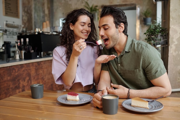 Young cheerful woman looking at her boyfriend eating piece of tasty cheesecake