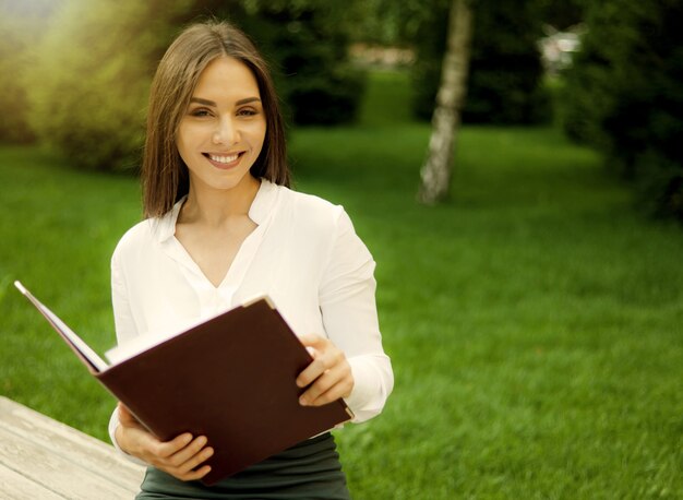 Молодая веселая женщина-менеджер держит в руках папку с документами, сидя на скамейке в парке