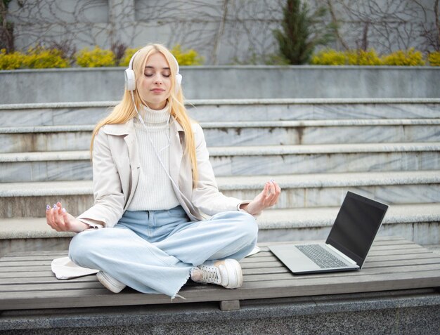 Photo young cheerful woman meditates in headphones while sitting on bench with a laptop