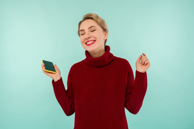 A young cheerful woman in a red sweater with a smartphone on a blue background